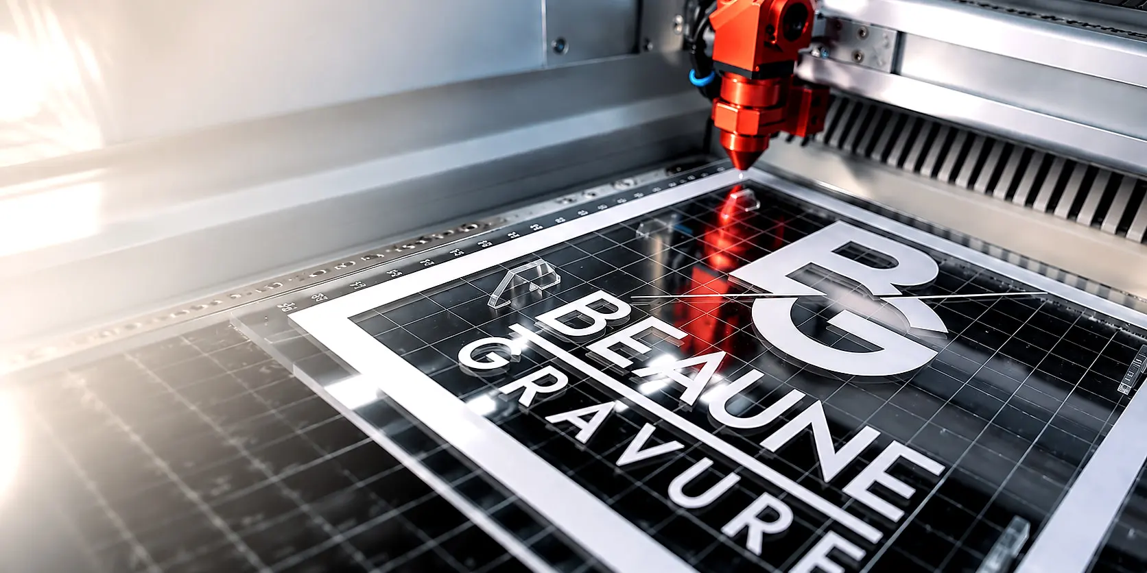 Tête de découpe laser rouge en action sur une plaque de plexiglass gravée au logo Beaune Gravure, dans l’atelier